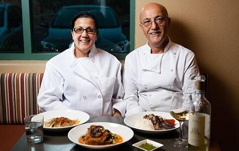 Saboor and Angela in white chef coats seated at a table of Mediterranean dishes and a bottle of white wine